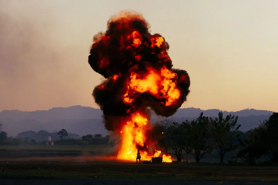A large explosion with fiery flames and thick smoke in an open field during sunset. Silhouetted figure nearby.