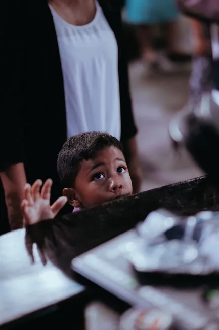 A curious child gazes upward from behind a counter indoors, reaching with a raised hand.