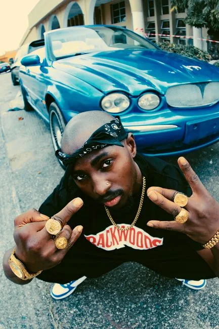 Man in gold jewelry and bandana poses crouching beside blue car in Lagos street.
