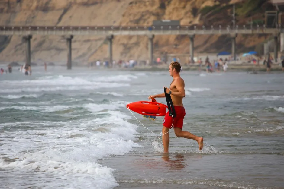 Lifeguard running into the ocean holding a rescue tube at a beach with a pier in the background.