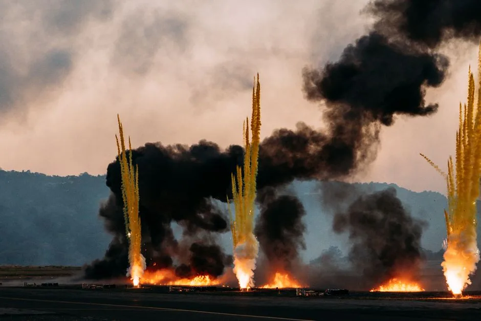 Intense scene of fire eruptions and smoke plumes rising against a cloudy sky, creating a dramatic atmosphere.