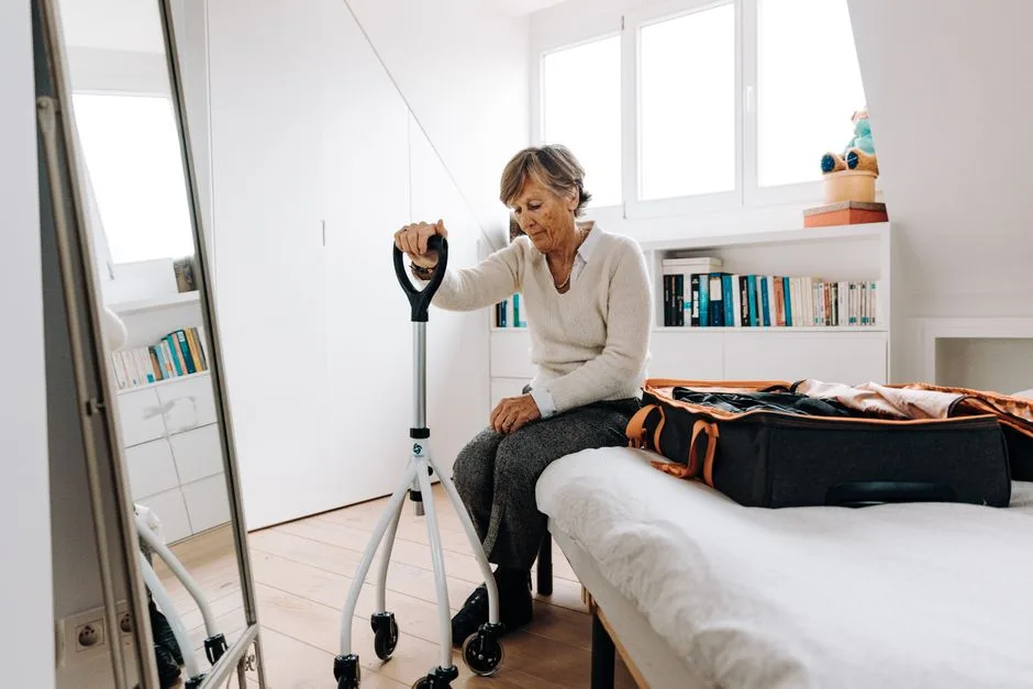 Senior woman sitting on bed with walker, preparing suitcase in bright bedroom.