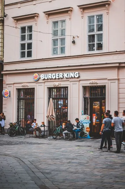 Street view of people sitting at tables outside a Burger King restaurant on a city pavement.