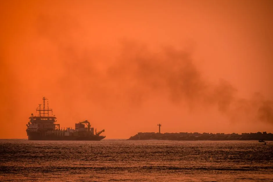 Silhouette of a ship sailing near the coastline during a vibrant orange sunset, emitting smoke against the horizon.