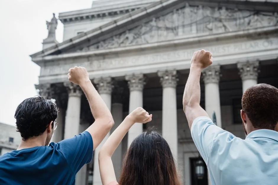 Three people raising fists in protest outside a government building with Greek-style architecture.