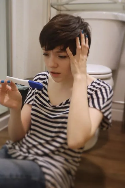 Young woman anxiously holding a pregnancy test while sitting on the bathroom floor.